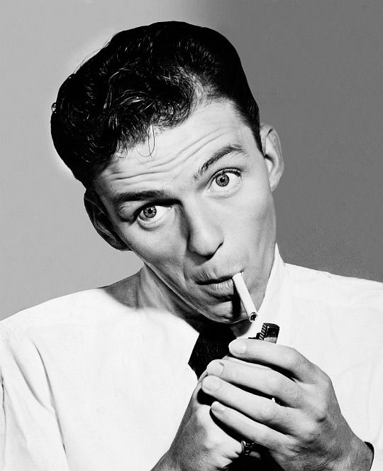 Black and white portrait of a young man with dark hair, wearing a light-colored shirt, and holding a lighter and a cigarette to his lips, with a serious expression.