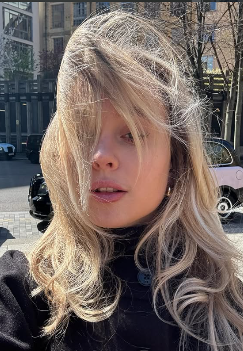 A woman with long blonde hair partially covering her face, standing outdoors on a sunny day.
