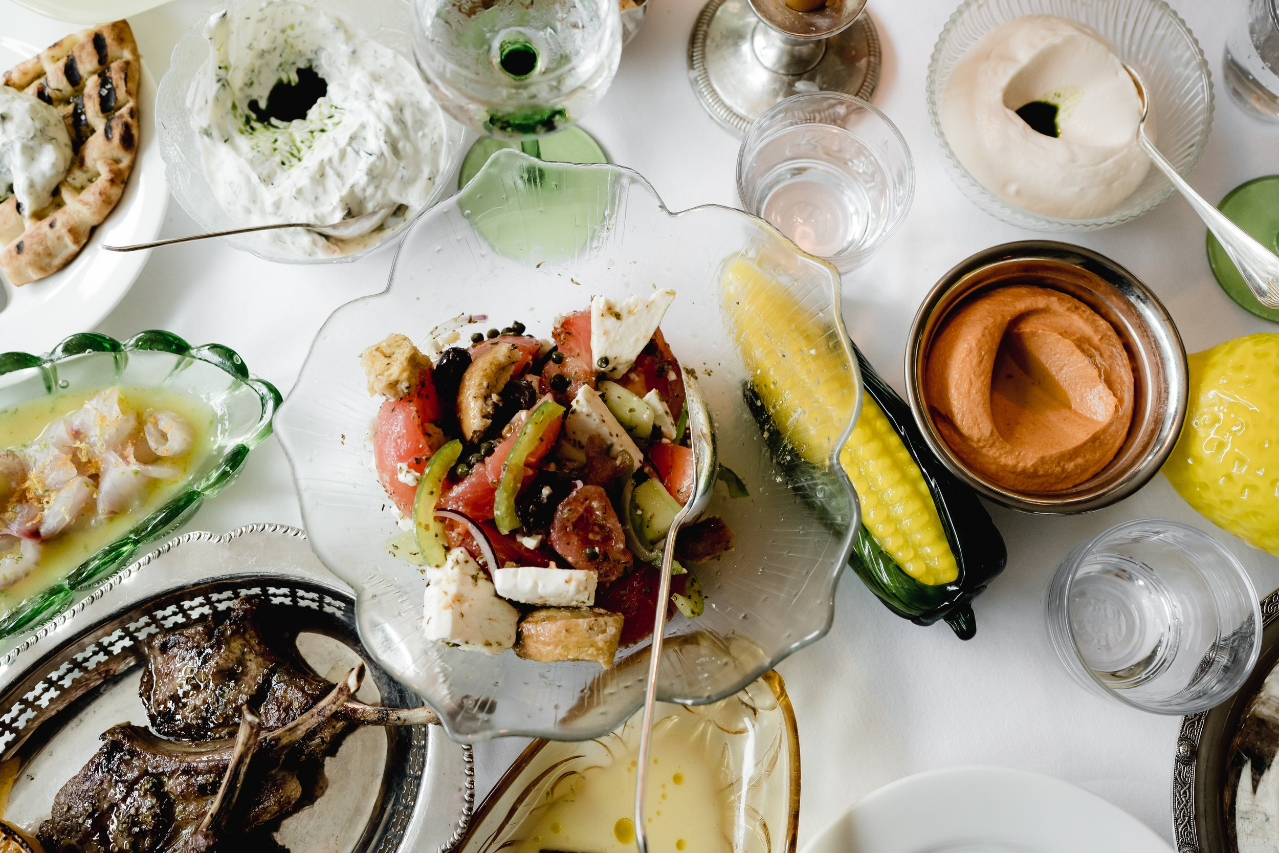 A variety of dishes on a table, including a Greek salad with tomatoes, cucumbers, and feta cheese, a bowl of hummus, a dish with grilled meat, various dips and sauces, and a glass of water.