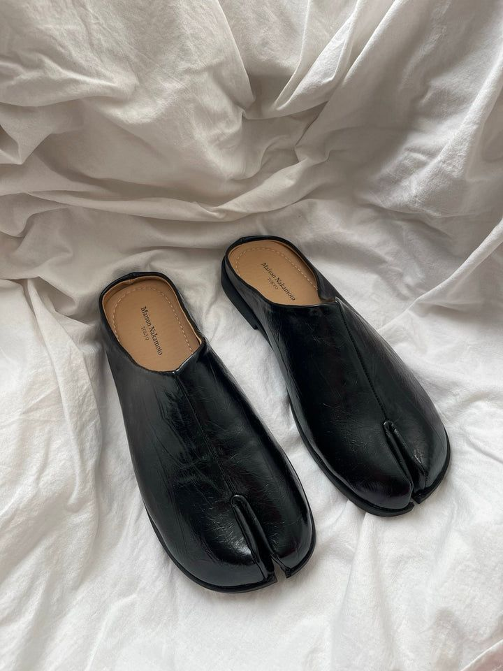 A pair of black leather slip-on shoes with a partial split toe design resting on white fabric.
