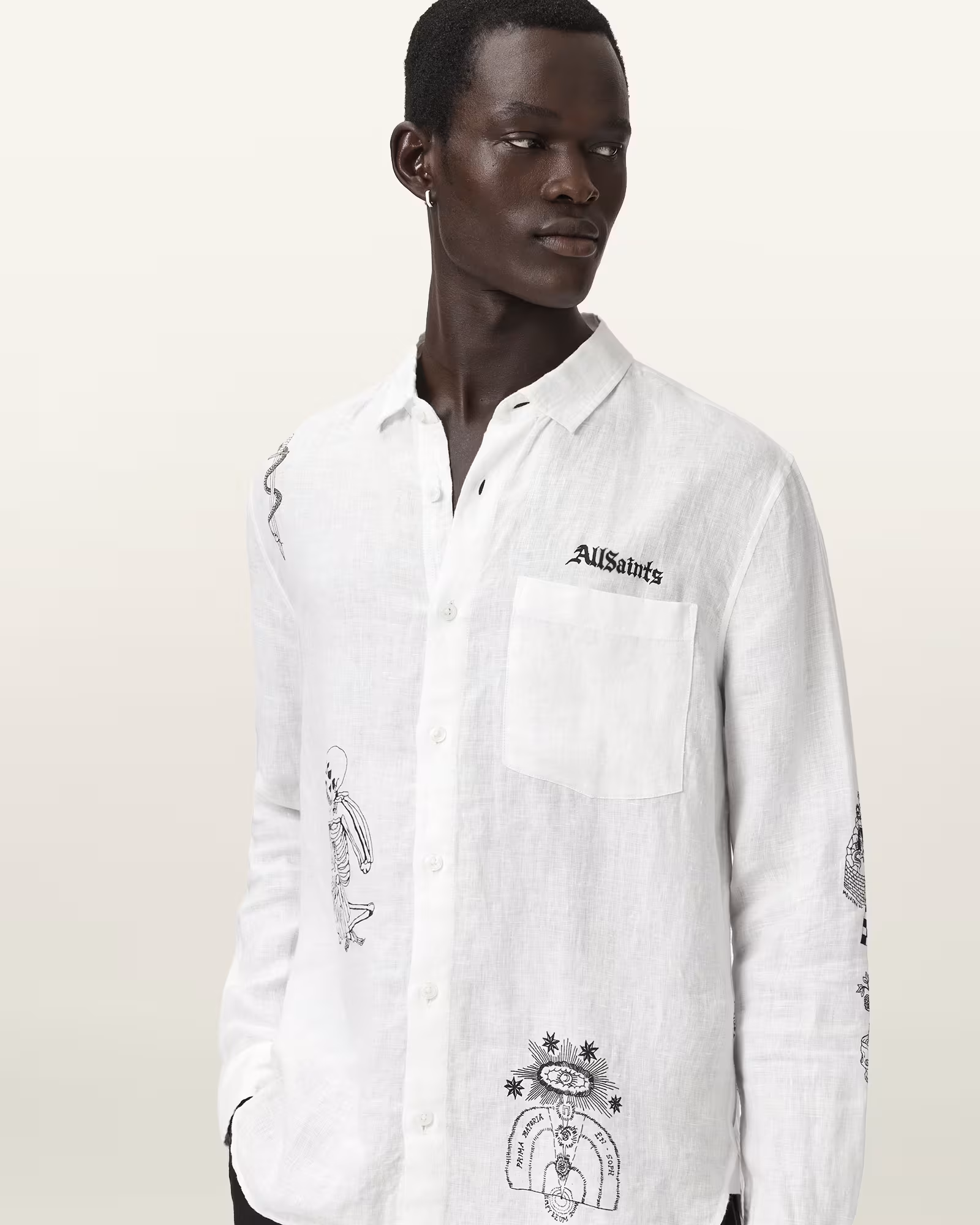 A dark-skinned man with short hair wearing a white button-up shirt with black artwork and text. The shirt features various illustrations, including a skeleton, a circular emblem, and other designs. The man is looking to his right against a plain light background.