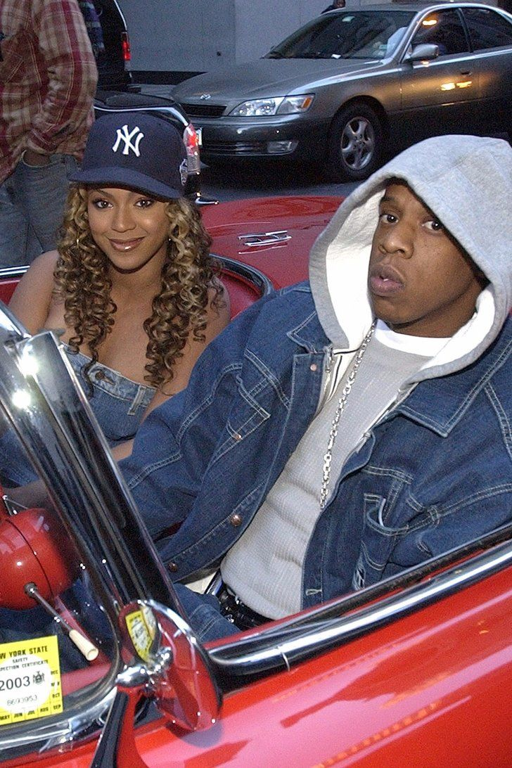 A woman with curly hair wearing a New York Yankees cap and a denim top, sitting in a red convertible car, smiling. A man wearing a gray hoodie, denim jacket, and chain necklace, sitting beside her, looking at the camera. There are parked cars and a person in a plaid jacket in the background.