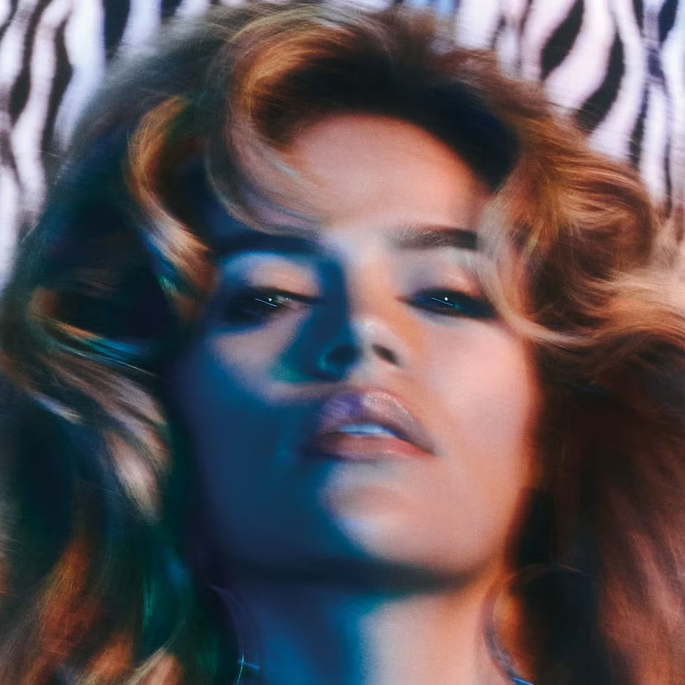 A woman with wavy blonde hair lying down with zebra print fabric in the background, looking at the camera with a sultry expression.