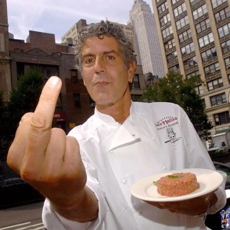 A chef wearing a white uniform with a logo on the chest holding a plate of raw meat and flipping off the camera with his middle finger against an urban city street background.