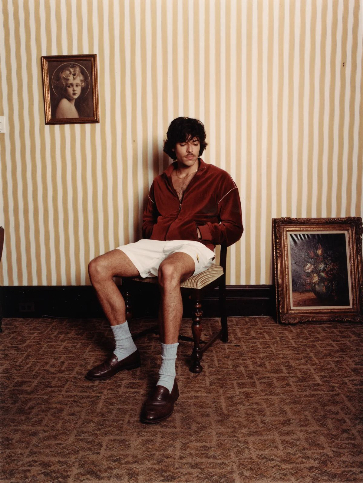 A young man with dark, curly hair sitting on a chair in a room with vintage striped wallpaper, wearing a red velour jacket, white shorts, white socks, and brown loafers. There are two framed artworks, one of a girl with curly hair on the wall and a floral still life on the floor.