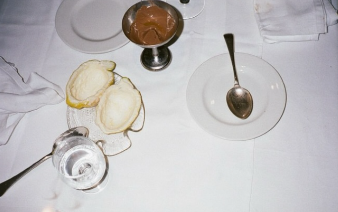 Empty white plate with a spoon, a glass of water, two lemon halves, a glass containing chocolate mousse, and a spoon on a white tablecloth.