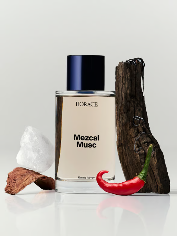 A perfume bottle labeled 'HORACE Mezcal Musc' surrounded by a chili pepper, a piece of wood, a rock, and a small piece of bark on a white surface with a plain background.
