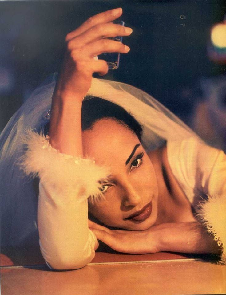 A woman lying on the floor with her head resting on her hand, gazing at the camera. She is wearing a white garment with fluffy detailing at the wrists and is holding a small glass in her raised hand above her head. She has dark hair pulled back, bold makeup, and expressive eyes, with a transparent veil draped over her head.