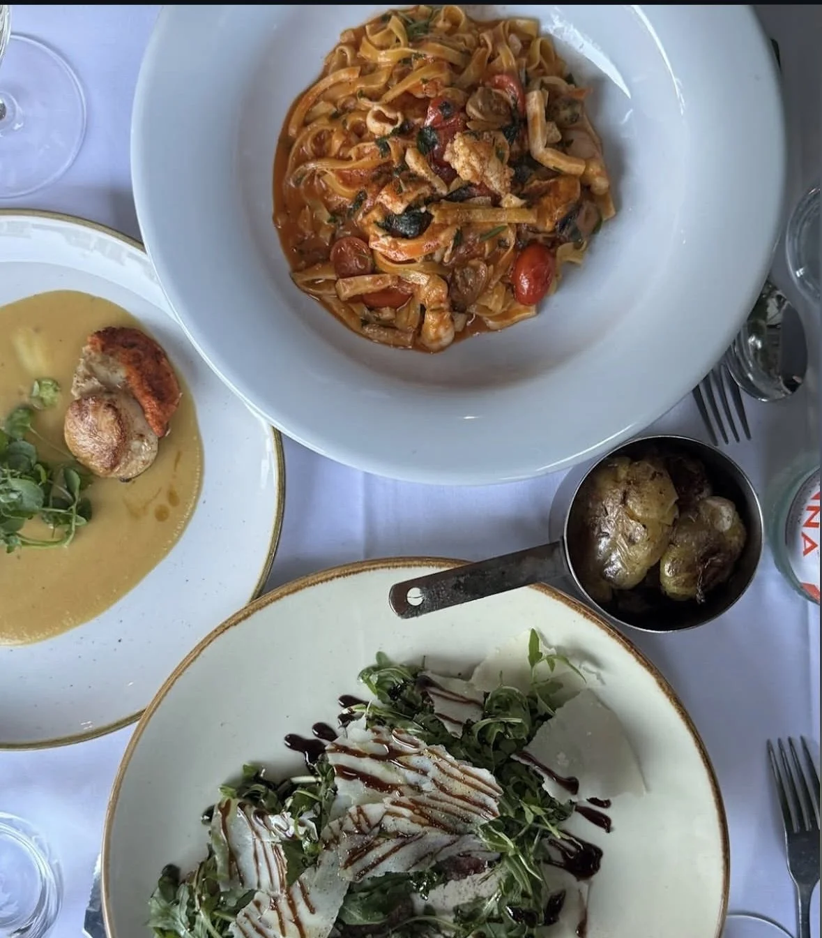 Assorted dishes including pasta with cherry tomatoes, herbs, and chicken, a salad with chicken and greens drizzled with balsamic reduction, and a bowl of potatoes with a fork on a white tablecloth.