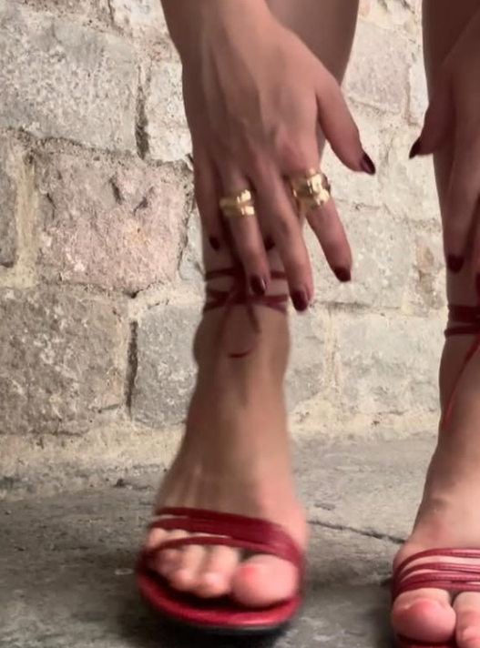 Close-up of a person's feet in red strappy heels, with hands adjusting the heel straps, jewelry on fingers, and a stone wall in the background.
