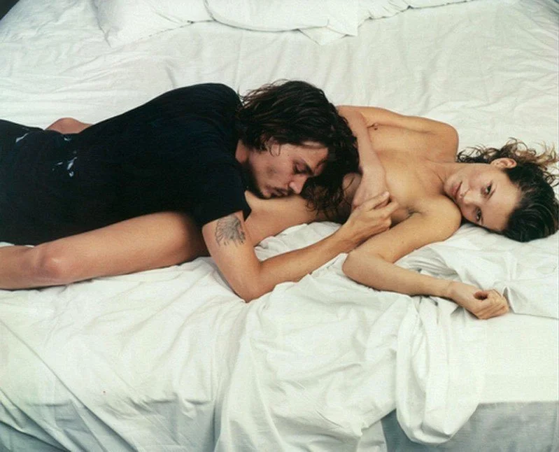 A man and woman lying on a bed, the man resting his head on the woman's chest, embracing her