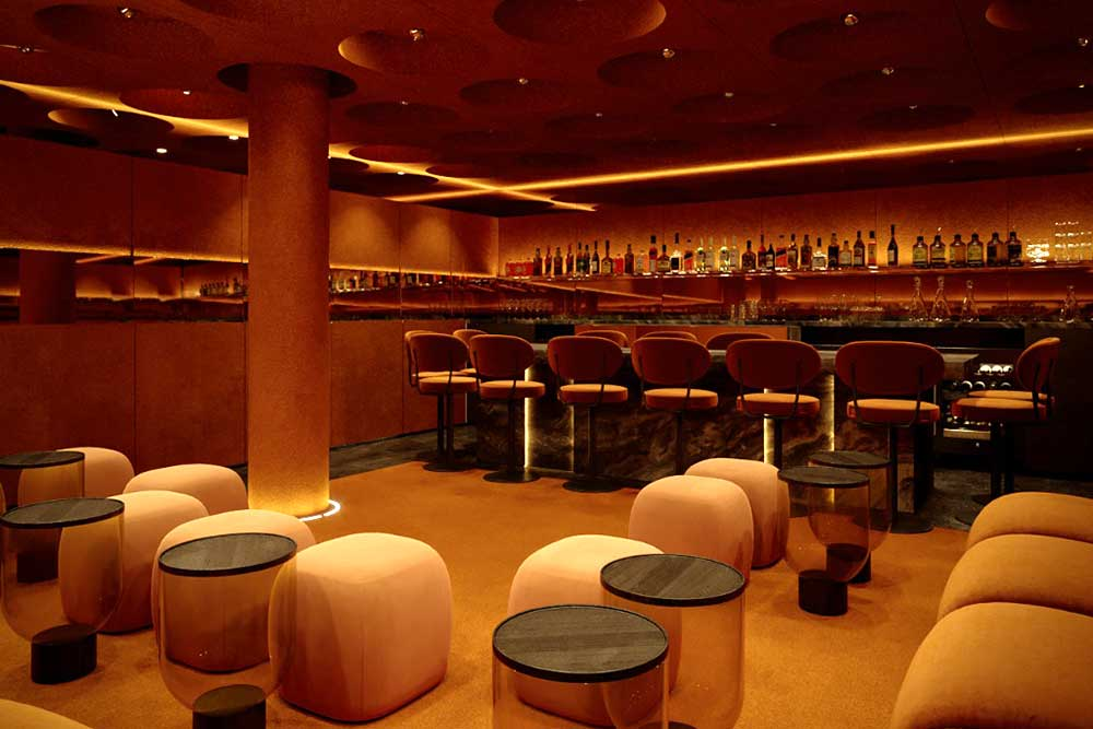 An empty upscale bar with orange and beige seating, small round black tables, and a well-stocked bar with various bottles in the background, illuminated by warm lighting.