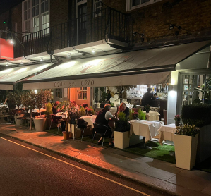Outdoor dining scene at night with tables and chairs, people dining, and warm lighting, in an urban setting.