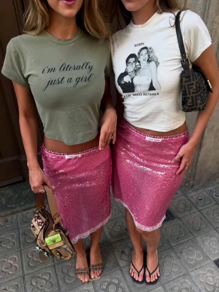 Two women standing side by side, wearing pink sequin skirts and cropped graphic t-shirts, with handbags and sandals. Their faces are partially out of the frame.