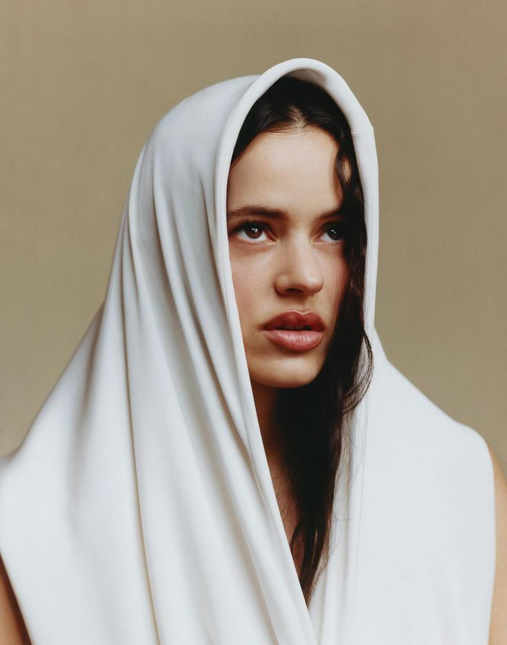 A woman with dark hair, wearing a white hooded garment, looks to the side against a neutral background.