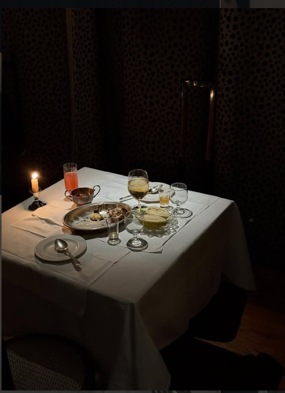 A dimly lit dinner table with a white tablecloth, set with various glasses, a candle, and leftover food.