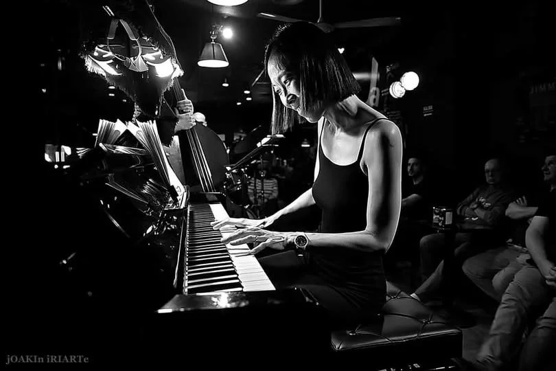 Black and white photo of a woman playing a grand piano in a dimly lit venue with audience members watching.