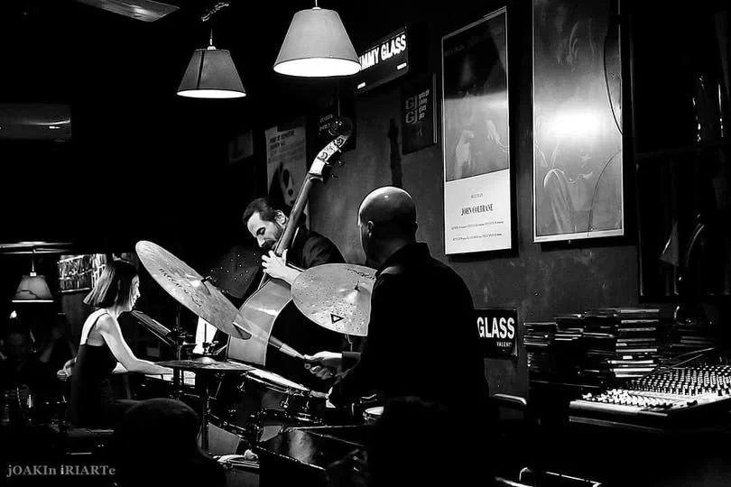 Jazz band performing live in a dimly lit club, featuring a guitarist, a bassist, and a drummer with a woman on piano or keyboard in the background.