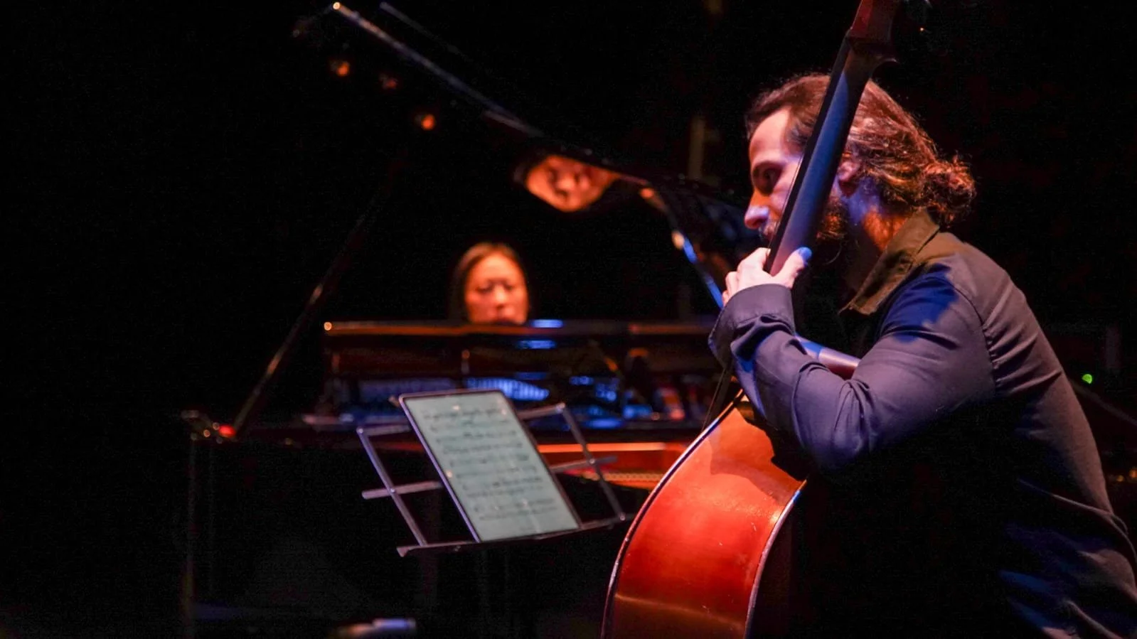 Two women playing musical instruments on a dark stage. One woman is playing a grand piano, and the other is playing a cello.