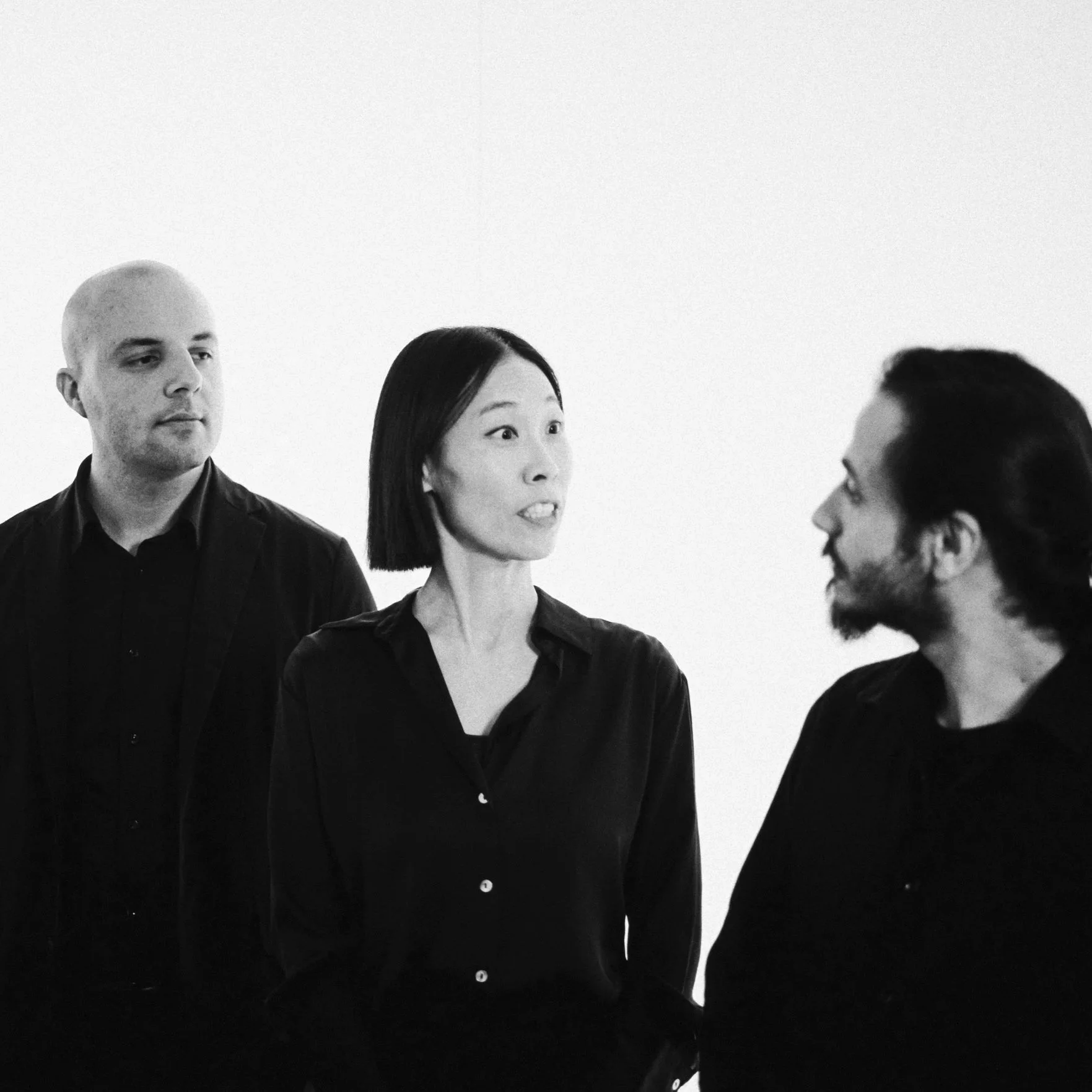 Three people having a conversation in a room, a woman in the center speaking to a man on the right, and a man on the left looking on, all dressed in black, against a plain light background.