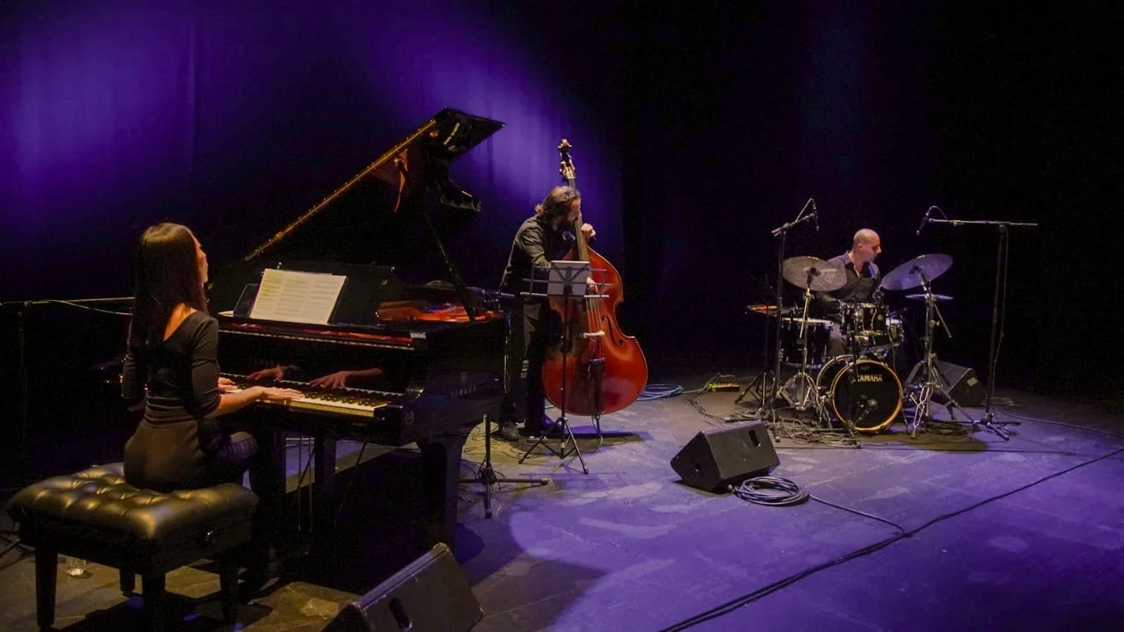 Jazz band performing on stage with piano, upright bass, and drums, under blue stage lighting.