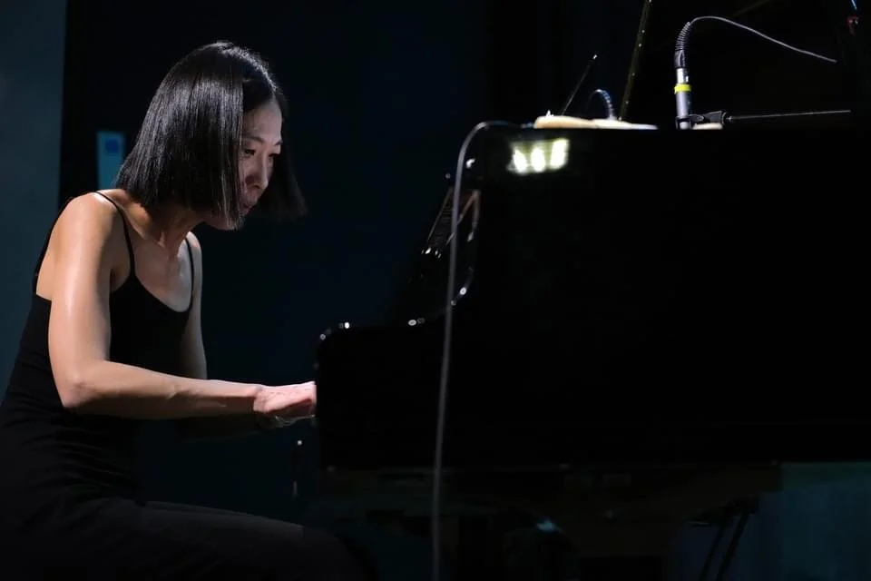 A woman with short black hair playing a grand piano in a dimly lit room