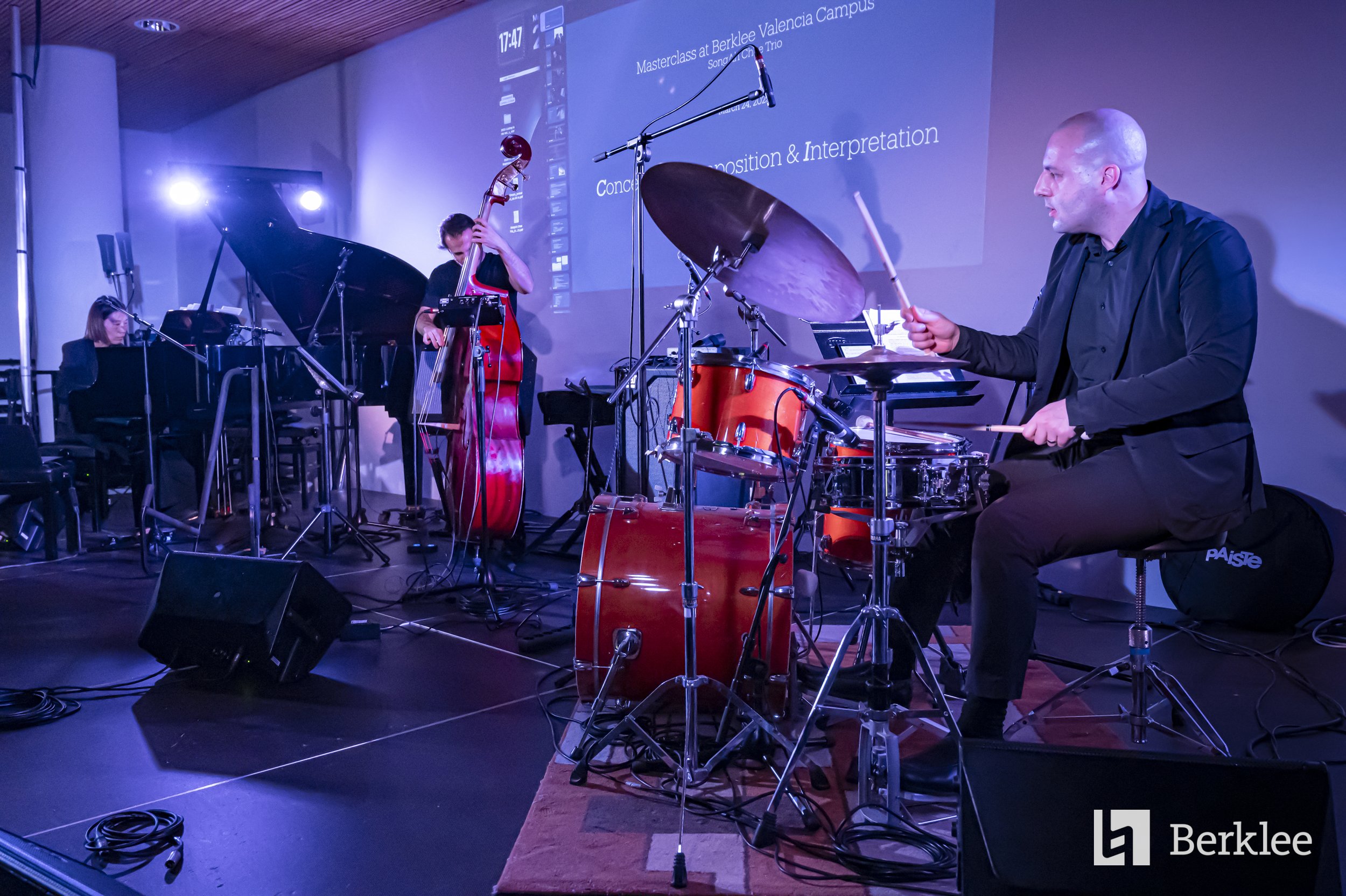 Jazz band performing on stage with a pianist, double bassist, and drummer, with a projection screen in the background displaying the event details, at Berklee Valencia Campus.