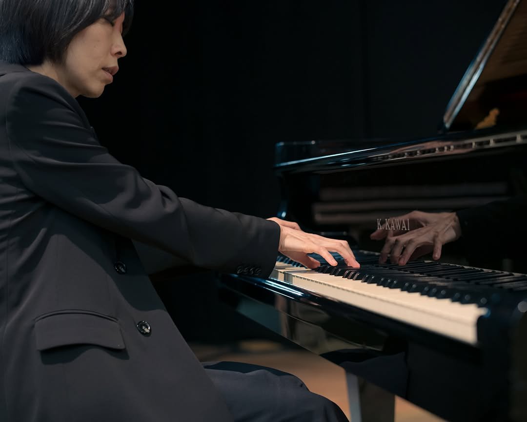 A woman with dark hair playing a grand piano, with her reflection visible on the shiny black surface of the piano.