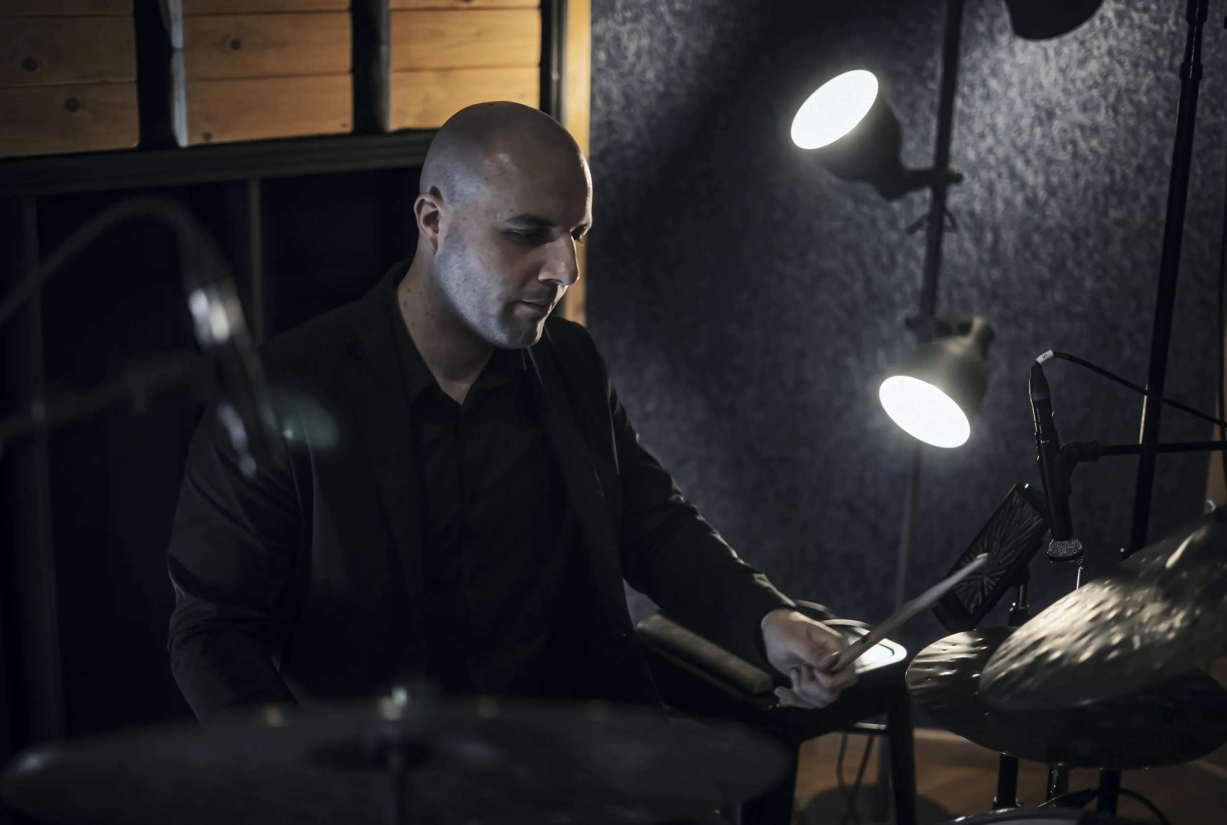 A bald man in a black shirt playing drums in a dimly lit recording studio.