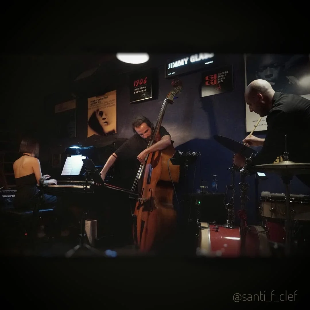 Jazz band performing on stage with three musicians: a woman playing piano, a man playing double bass, and another man on percussion with a drum set, in a dimly lit venue.