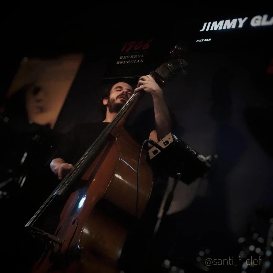 A man playing an upright bass on stage at a jazz bar, with signs for "Jimmy G" and others visible in the background.