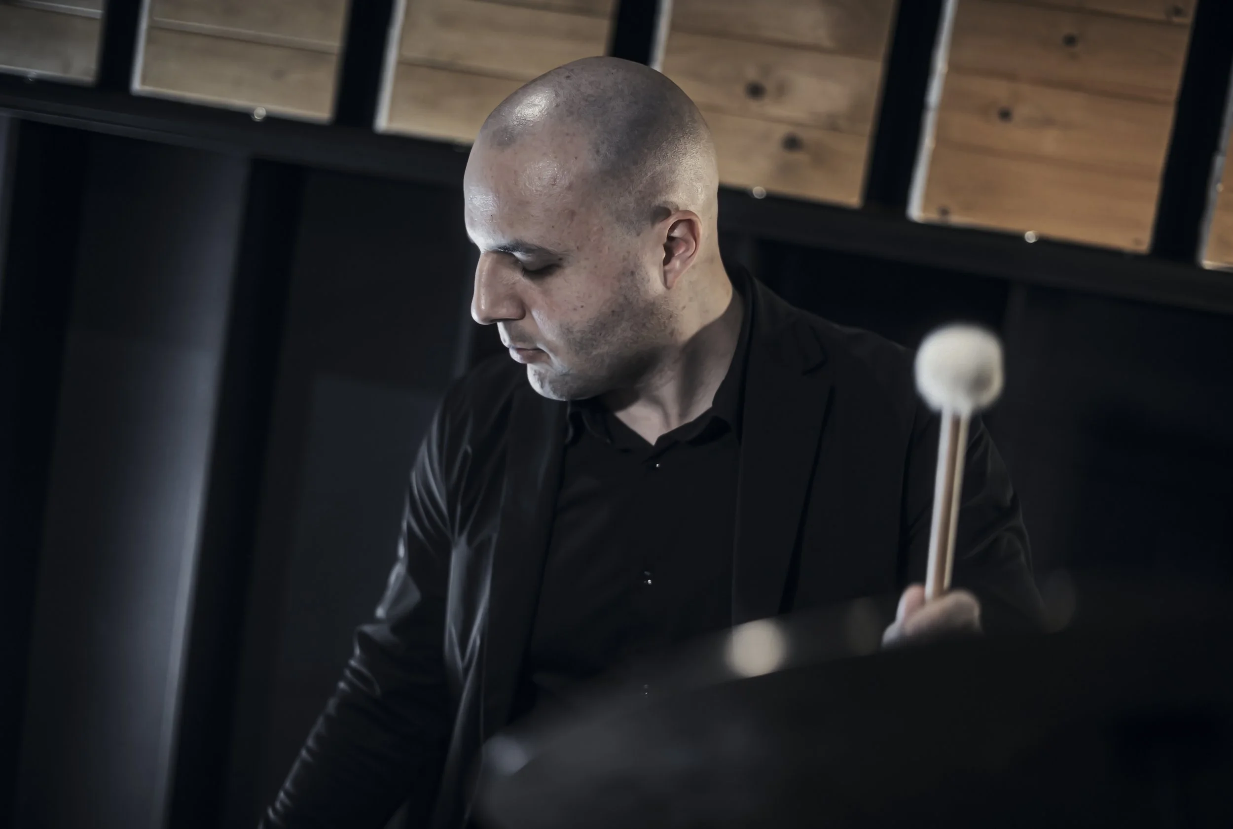 A man with a shaved head wearing a black suit in a dark room with wooden paneling, looking down as he holds what appears to be a mallet.