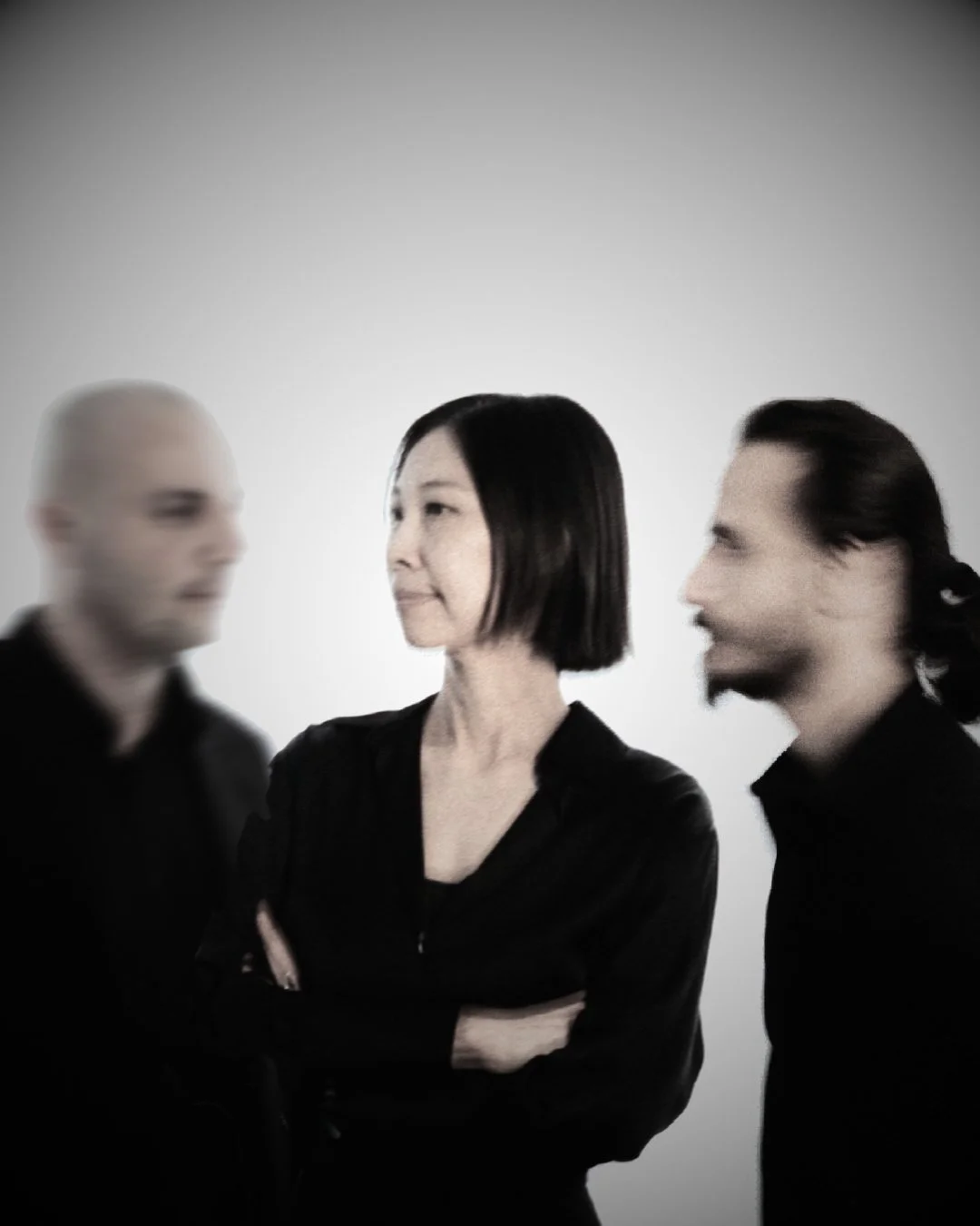 Three people, one woman and two men, standing against a light gray background. The woman is centered with black hair styled in a bob, arms crossed, wearing a black top. The man on the left is blurred and bald, and the man on the right has long dark h