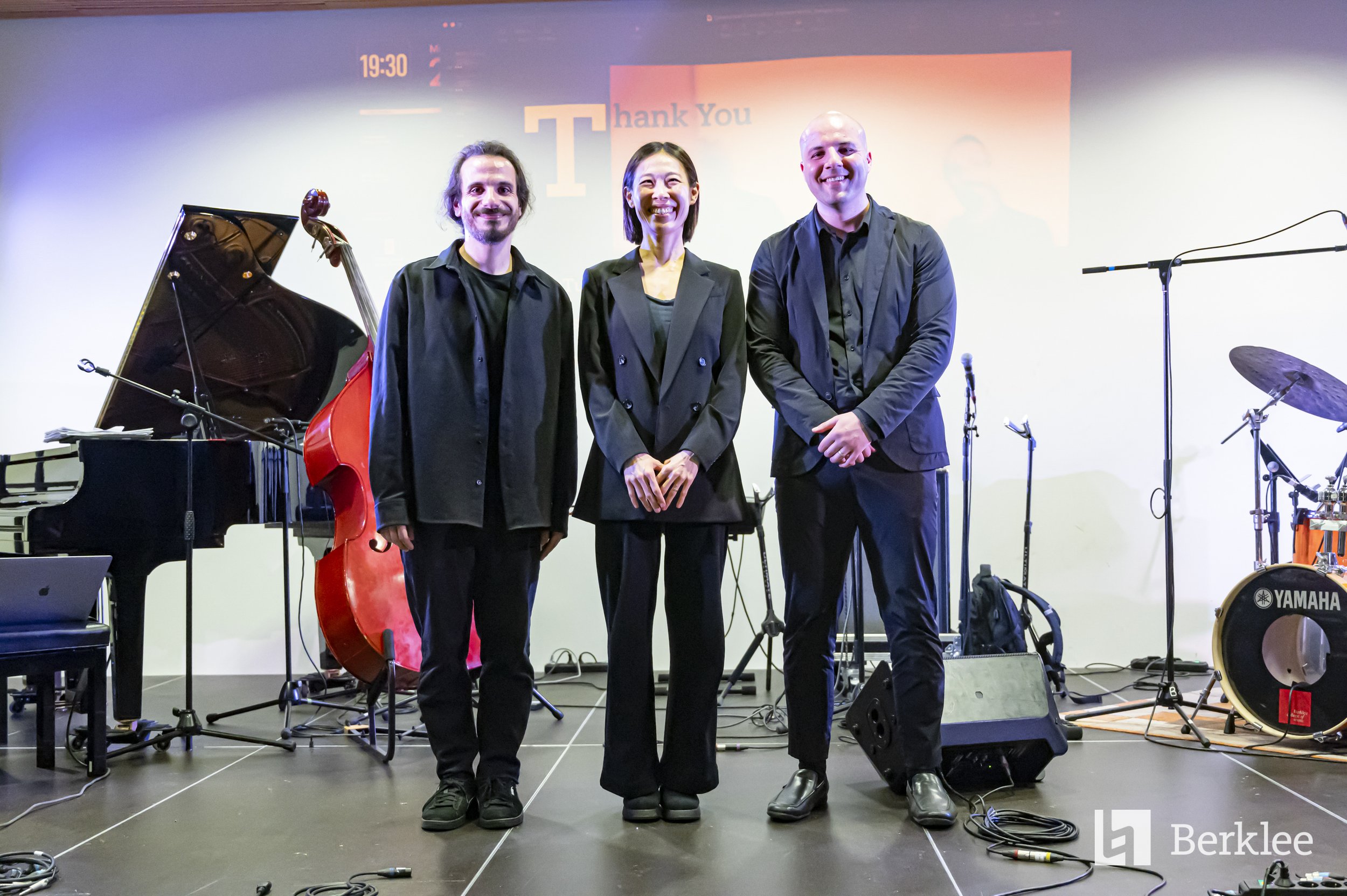 Three people standing on a stage with musical instruments behind them, smiling, in a setting that appears to be an event or concert, with a screen in the background displaying a 'Thank You' message.