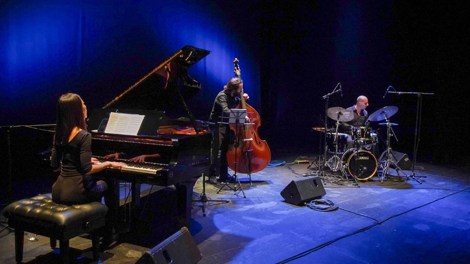 Jazz band performing on stage with piano, upright bass, and drums, under blue stage lighting.