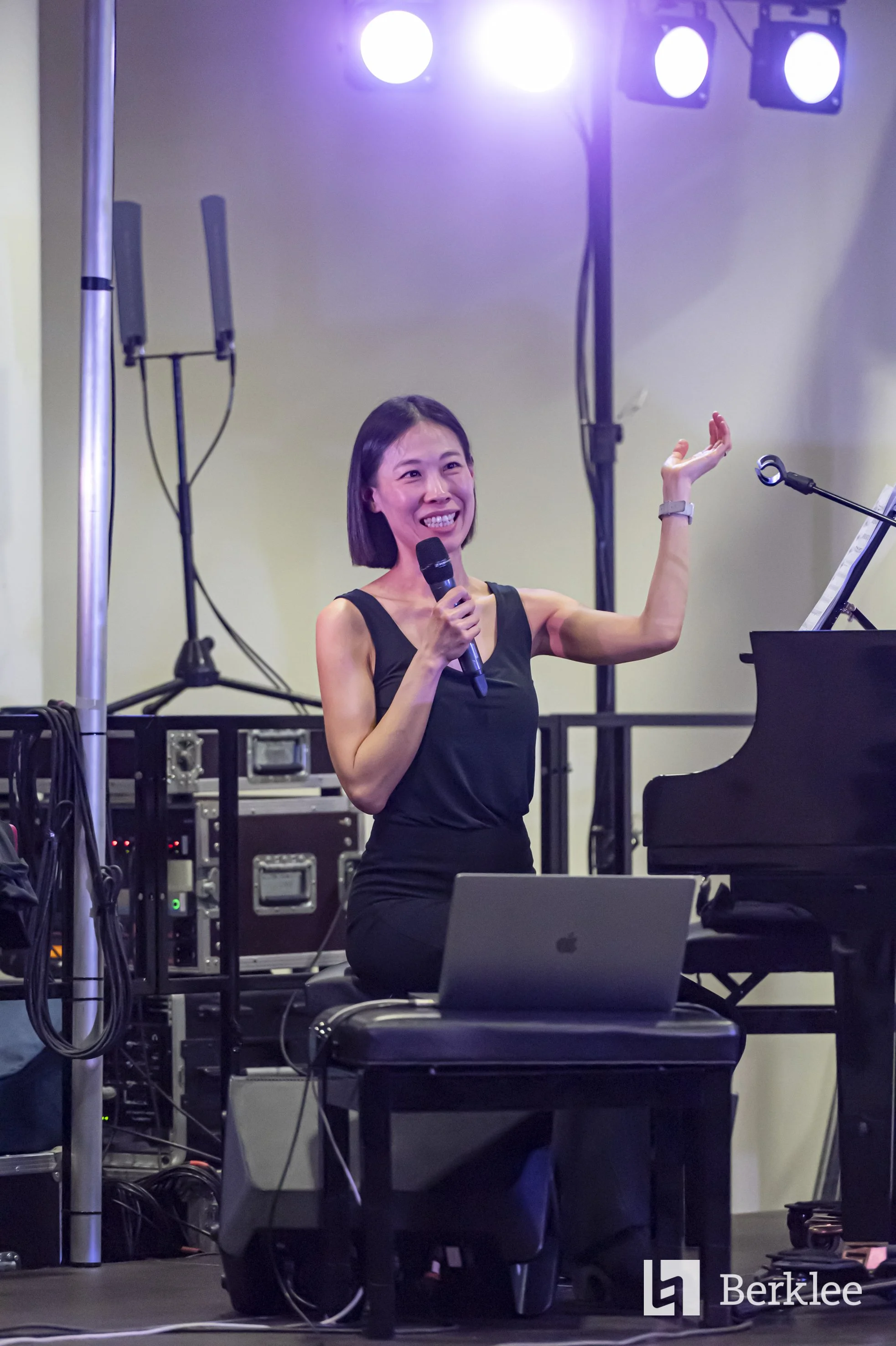 A woman in a black sleeveless dress holding a microphone and smiling during a performance, surrounded by stage equipment and a piano, with purple stage lights overhead.