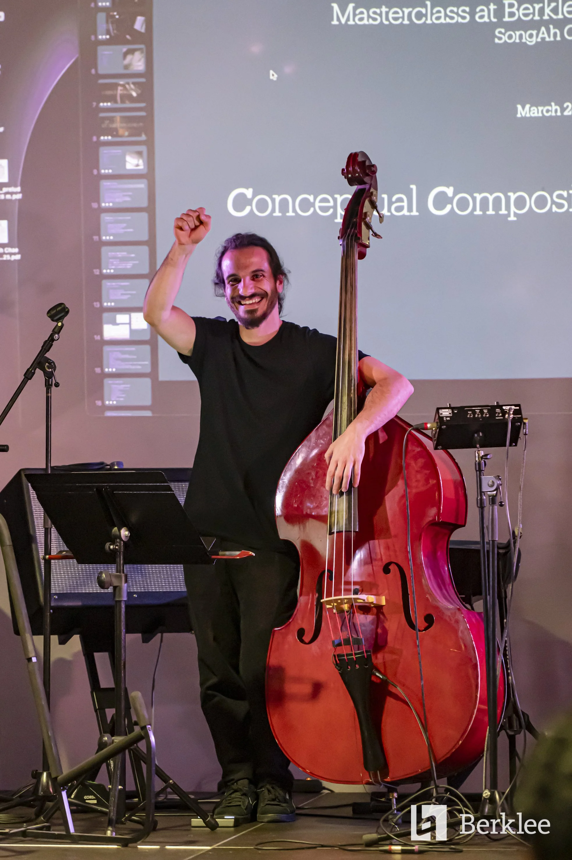 A man with long hair and a beard smiling and raising his fist, standing next to a double bass on a stage during a concert or event.