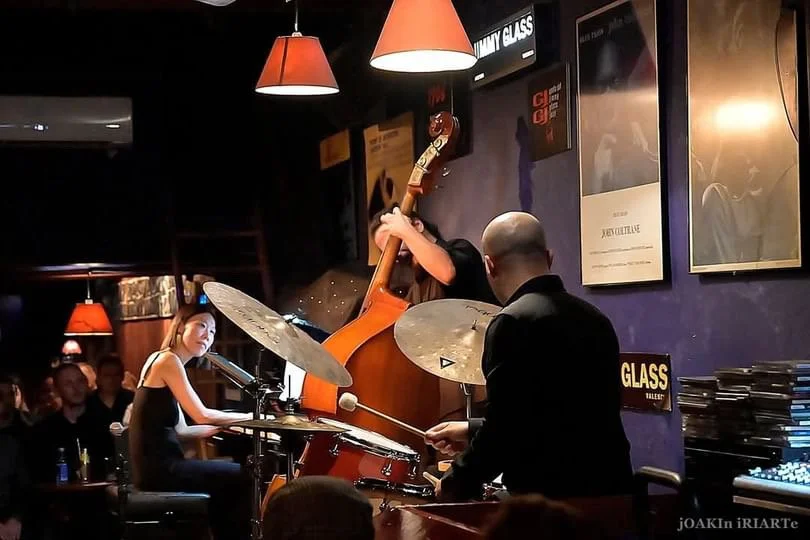 A jazz band performing live in a dimly lit bar, featuring a female pianist, a male drummer, and a double bass player.