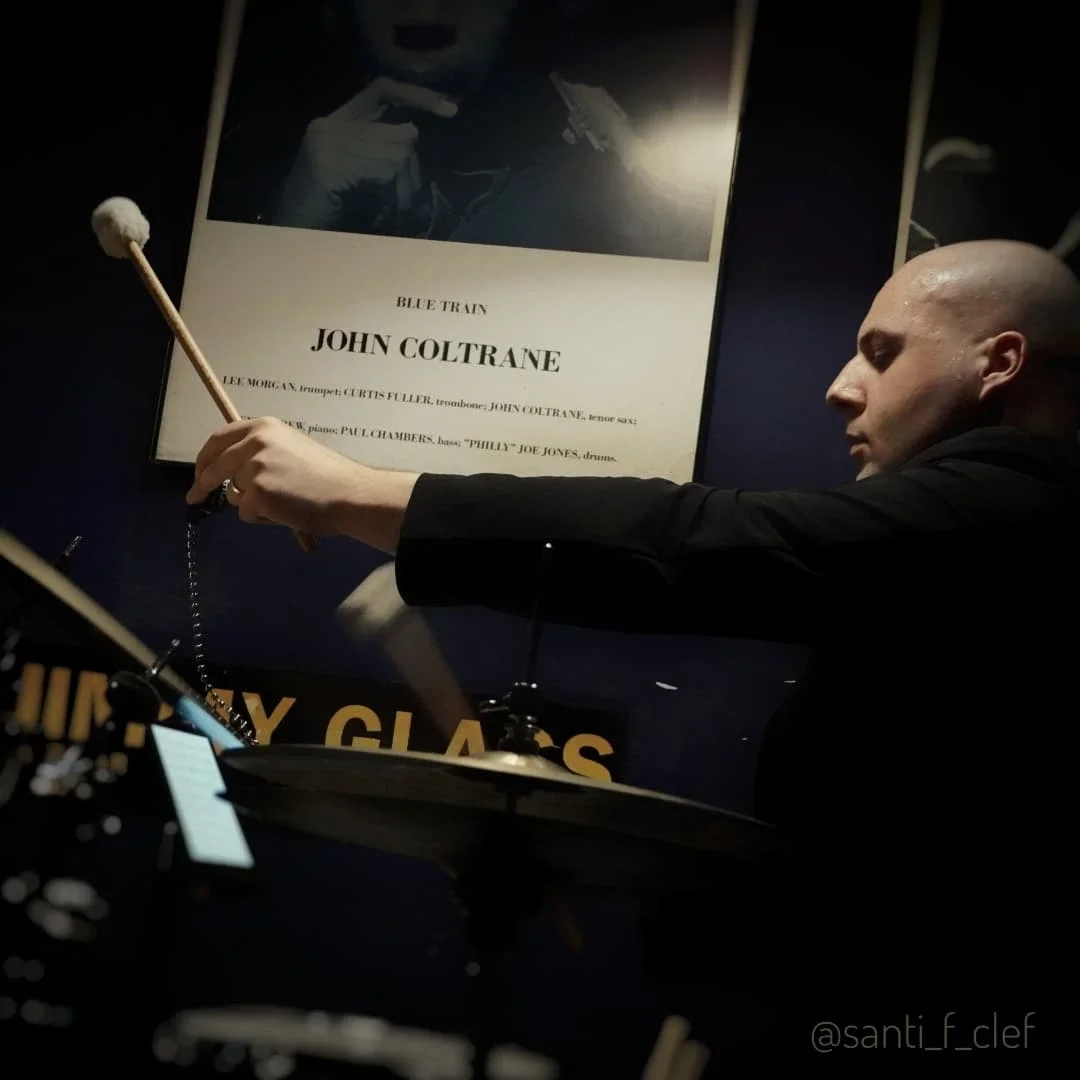 A man with a shaved head playing drums, with a poster of John Coltrane's album 'Blue Train' on the wall behind him.