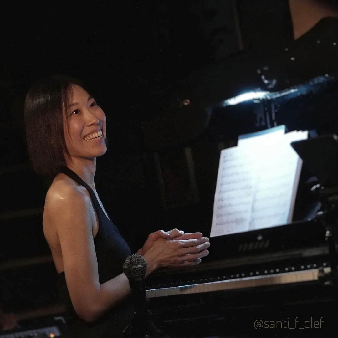 A woman smiling at a piano with sheet music, holding her hands together near her face, in a dark room.
