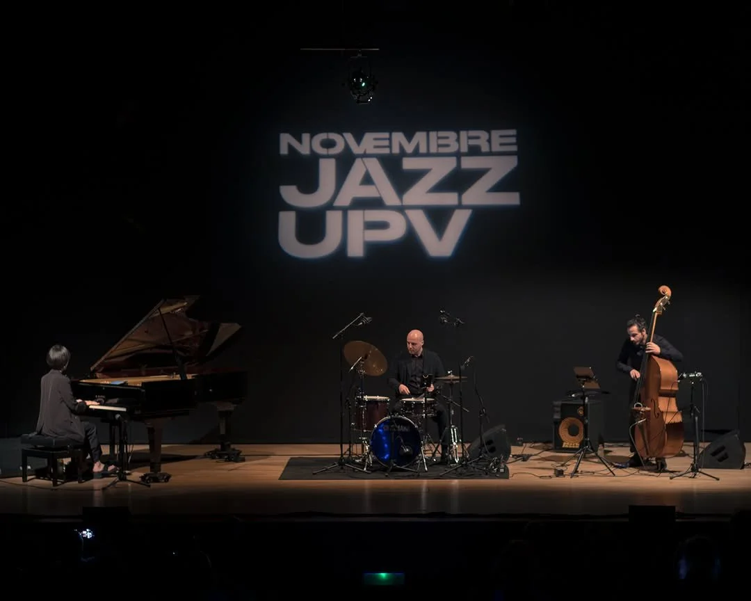 A jazz band performing on stage with a pianist, drummer, and double bassist, under a sign that reads 'Novembre Jazz UPV' in large letters.