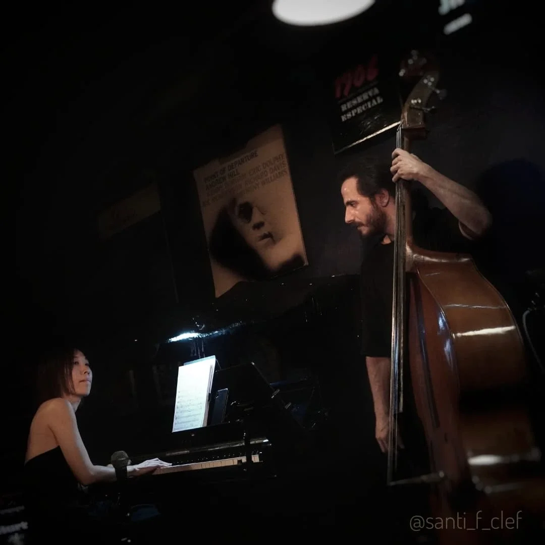 A woman playing a grand piano and a man standing beside her playing an upright bass in a dimly lit room with posters on the wall.