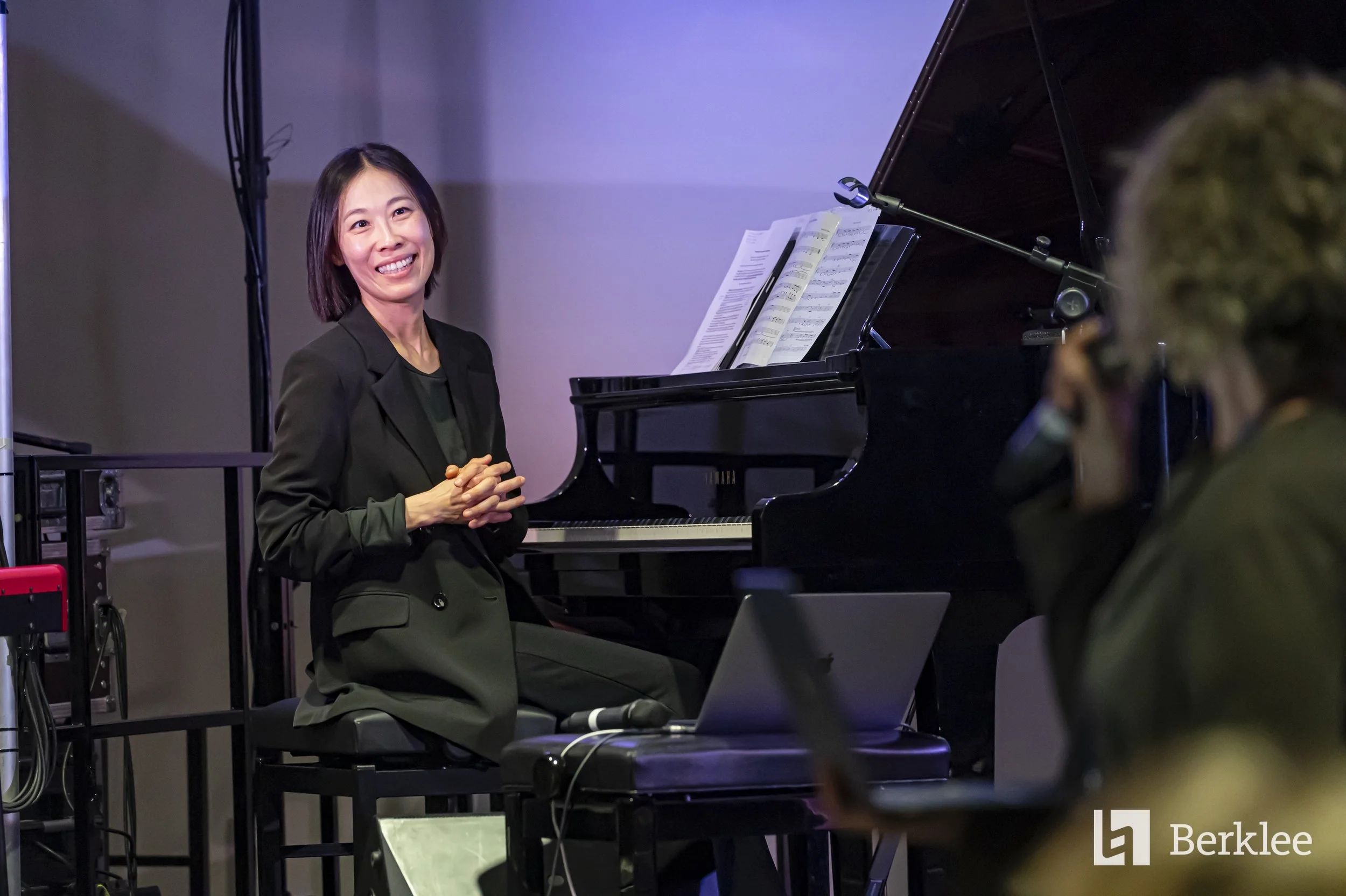 A woman sitting at a grand piano smiling and looking at a person with a headset who is taking a photo or recording. The woman has short dark hair and is wearing a black suit. There is a laptop and a microphone on a stand nearby.