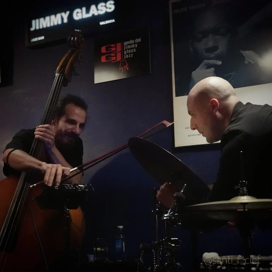 Two musicians perform live; one playing upright bass and the other on drums. There are black and white posters on the dark wall behind them.