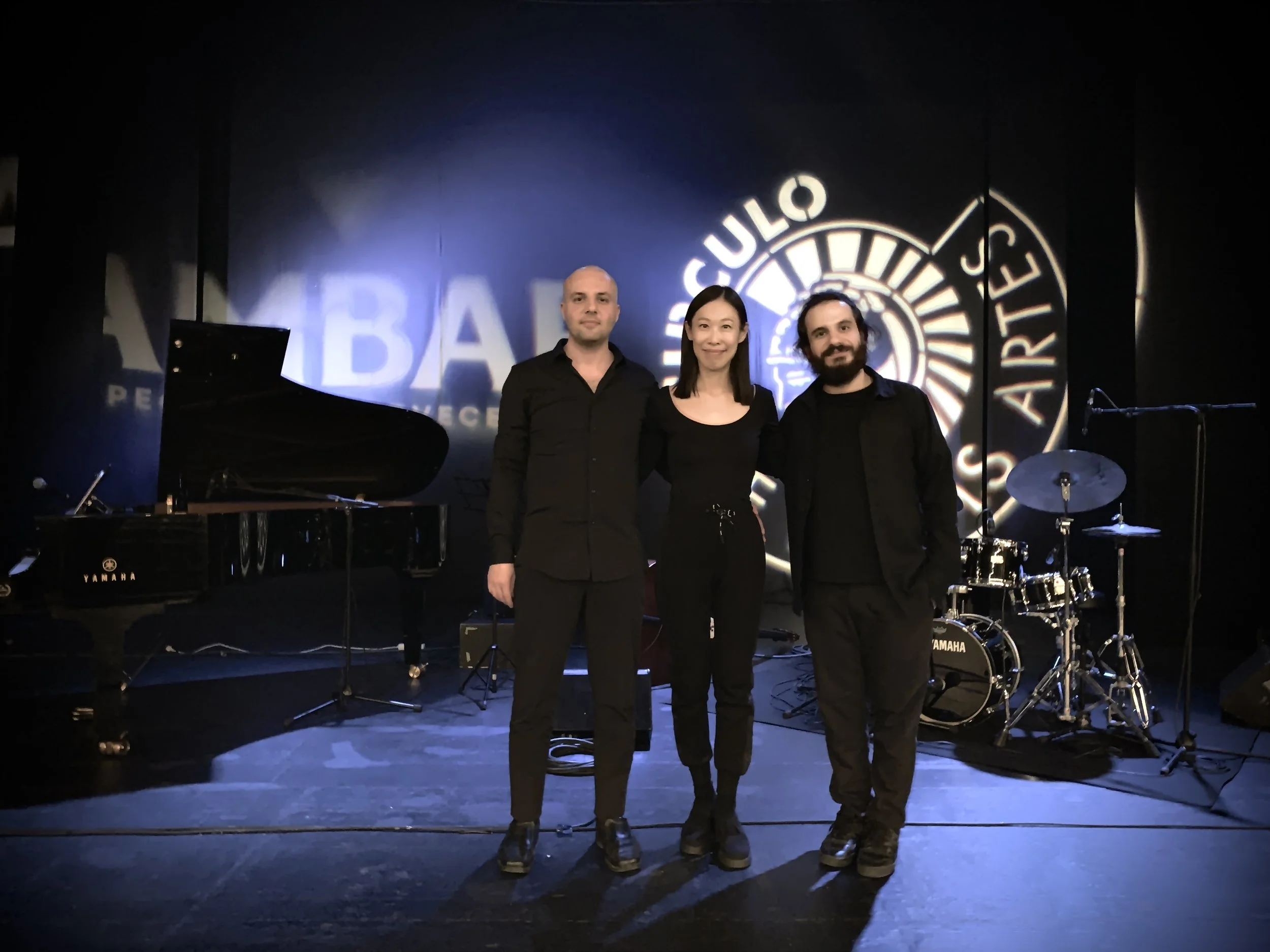 Three musicians stand on stage in front of a dark backdrop with a logo. The stage has a grand piano, drums, and microphone stands. The musicians are dressed in black, two men and one woman, smiling at the camera.