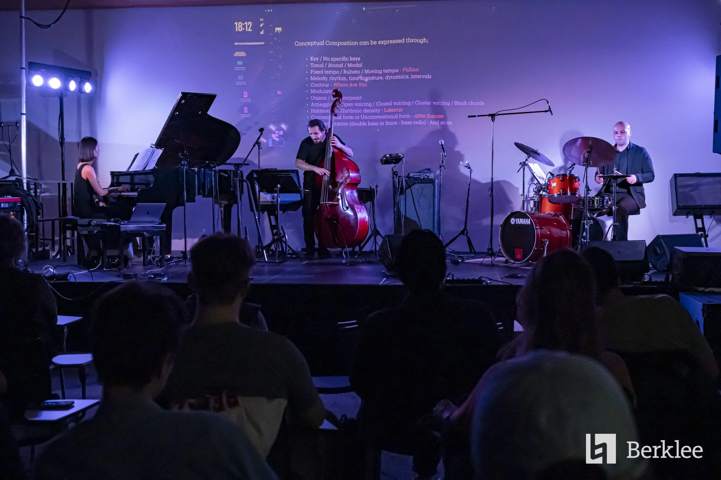 Musicians performing on stage with a pianist, double bassist, and drummer, while audience members watch in a dimly lit venue. A large screen behind displays text about musical concepts.