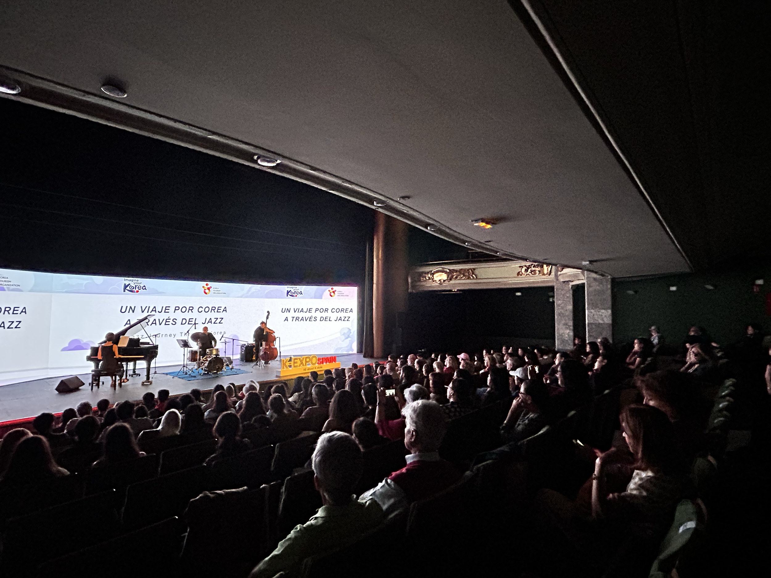 Audience watching jazz performance on stage with three musicians, piano, drums, and double bass, at an indoor event, with a large screen displaying text in Spanish and French.
