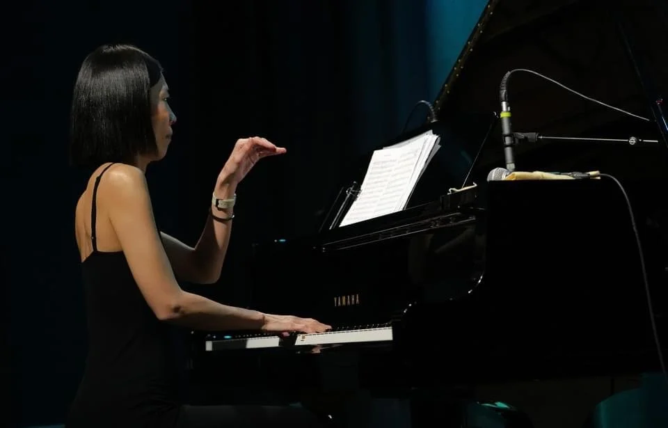 A woman with black hair playing a grand piano on stage with a music sheet in front of her.