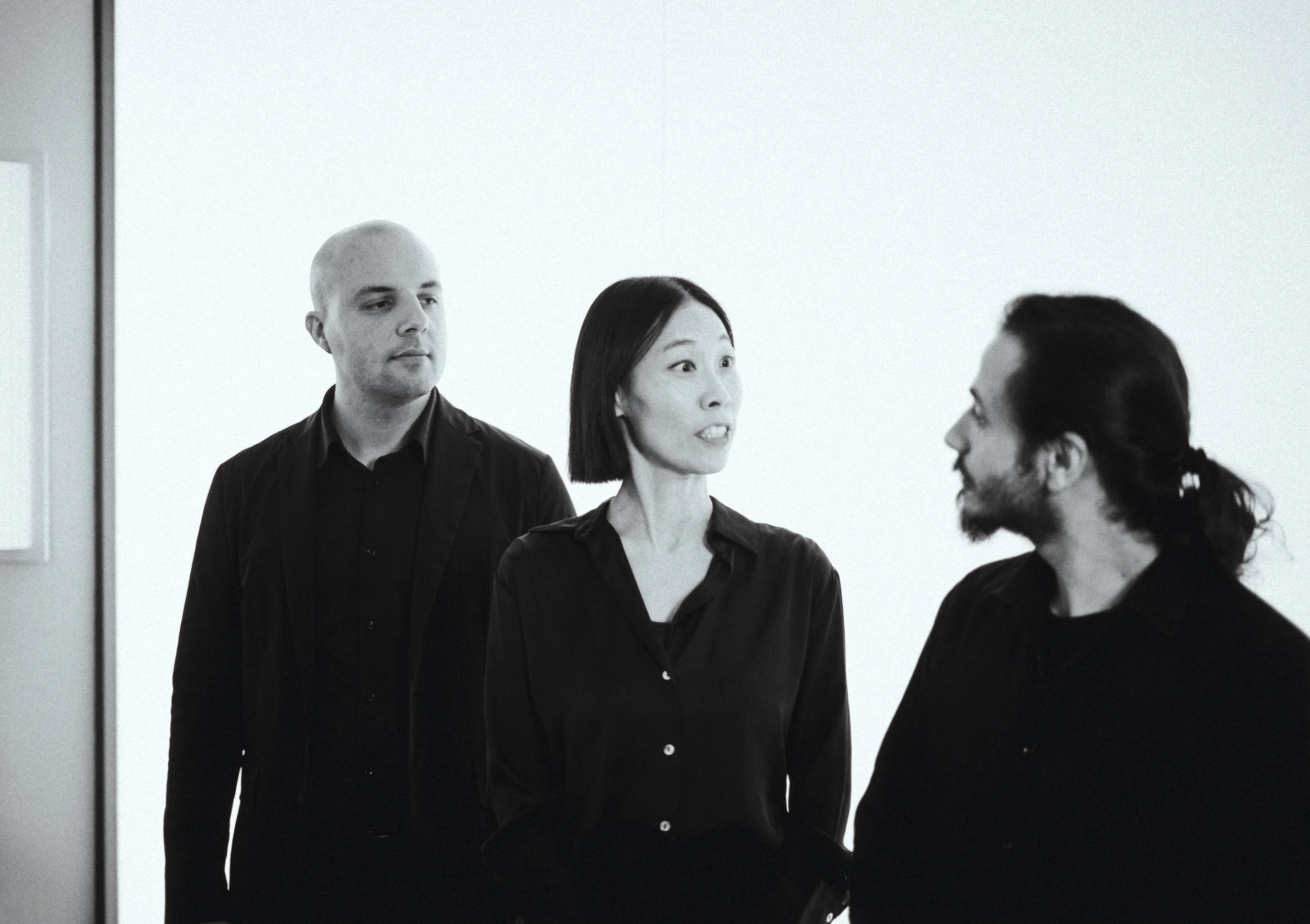 Three people having a conversation in a room, a woman in the center speaking to a man on the right, and a man on the left looking on, all dressed in black, against a plain light background.