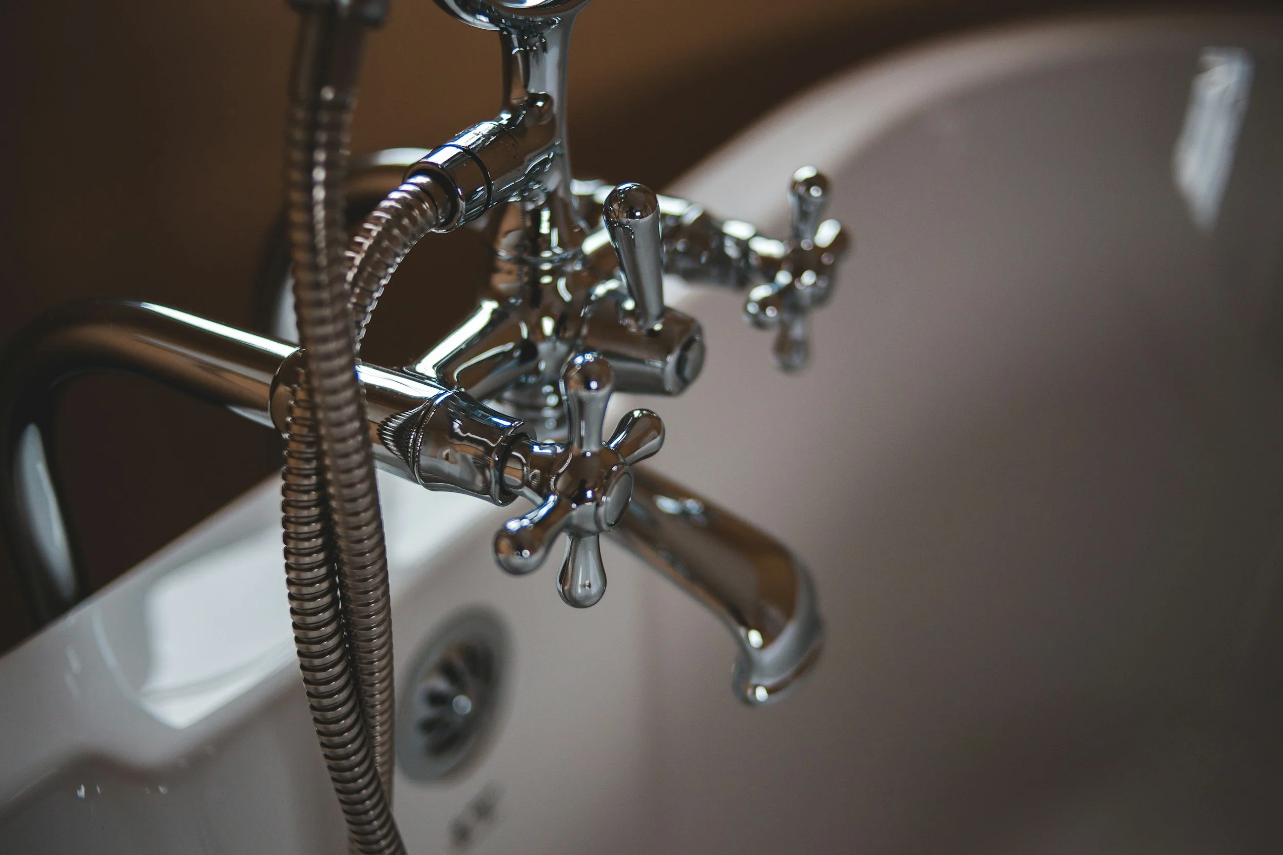 Close-up of a vintage-style chrome bathtub faucet with cross handles and metal hose attachment, mounted above a white bathtub.